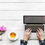 Hands typing on a laptop with flowers and a cup of tea nearby